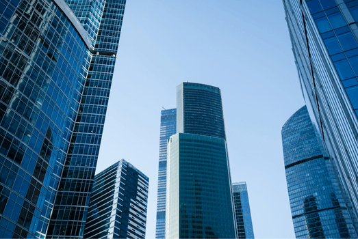 Modern glass skyscrapers rise against a clear blue sky, creating a sleek, imposing cityscape that conveys progress, ambition, and energy.