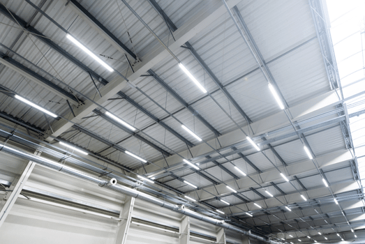 Bright industrial warehouse ceiling with exposed steel beams, metal roofing, ducts, and rows of fluorescent lights, creating a clean, modern tone. energy efficiency for operating companies