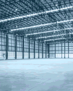 Vast empty industrial warehouse with steel roof trusses, bright strip lights, tall windows, and a smooth concrete floor in cool blue tones.
